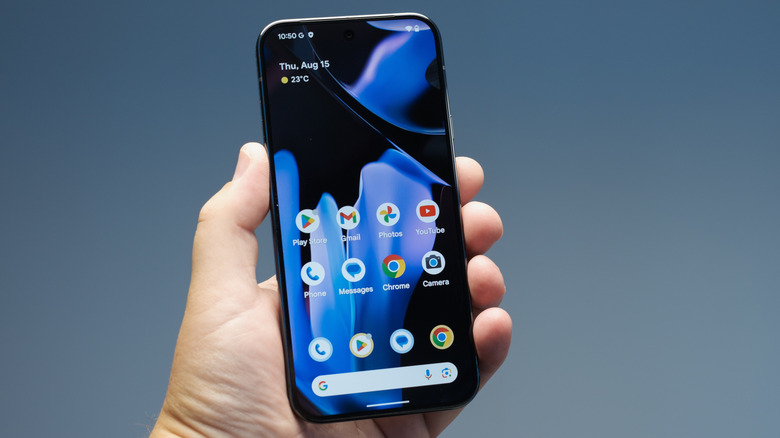 A black Google Pixel 9 held in an individual's left hand, the Pixel is displaying its home screen