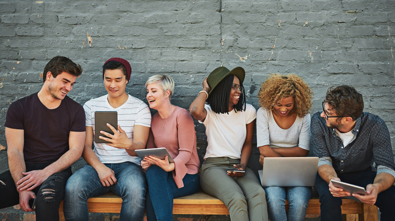 A group of people sitting on a bench laughing and holding different devices