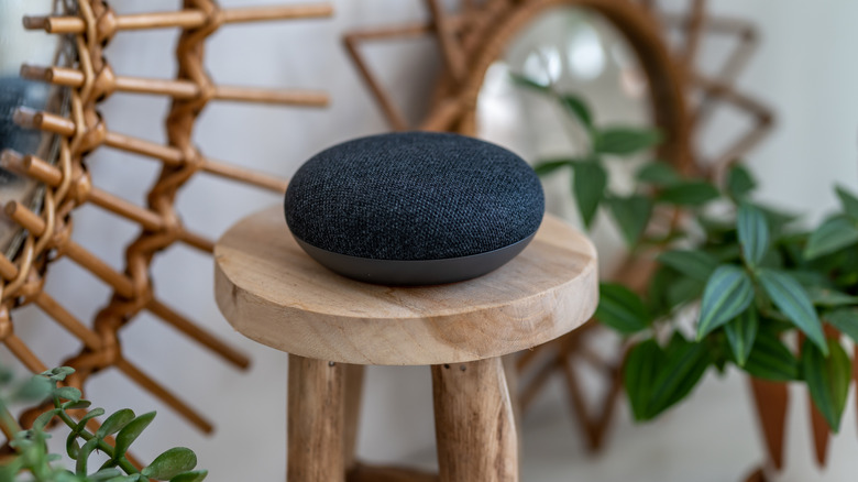 A black Google Home speaker sitting on a wooden stool, there are some plants surrounding it along with some wooden decorations on the wall behind it