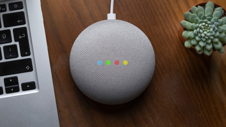 A grey Google Home assistant sitting between a silver laptop and a small cactus, the Google Home is displaying four colored dots in its center