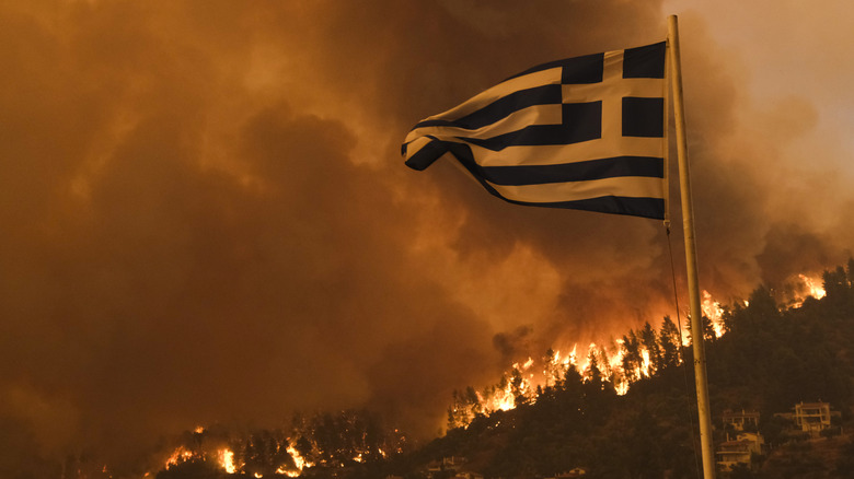 The Greek national flag flying in the wind with a wildfire burning in the background.