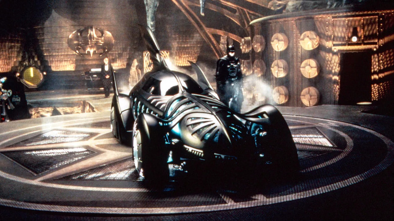 Batman standing next to his Batmobile in the cave with Alfred in the background