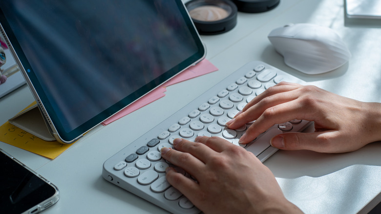 Close-up of hands typing on a compact white keyboard next to a tablet on a stand, with a wireless mouse and desk items in the background.