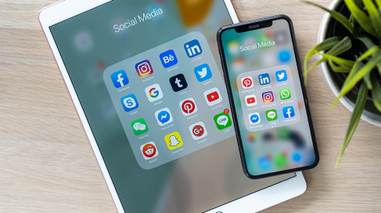 A tablet and smartphone lying on a desk display folders of social media apps, including familiar platform icons arranged on their screens, with a plant nearby.