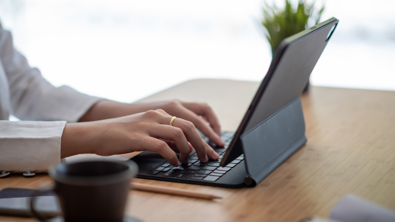 Person typing on a tablet with a keyboard case on a wooden desk, with a coffee cup and pencil nearby.