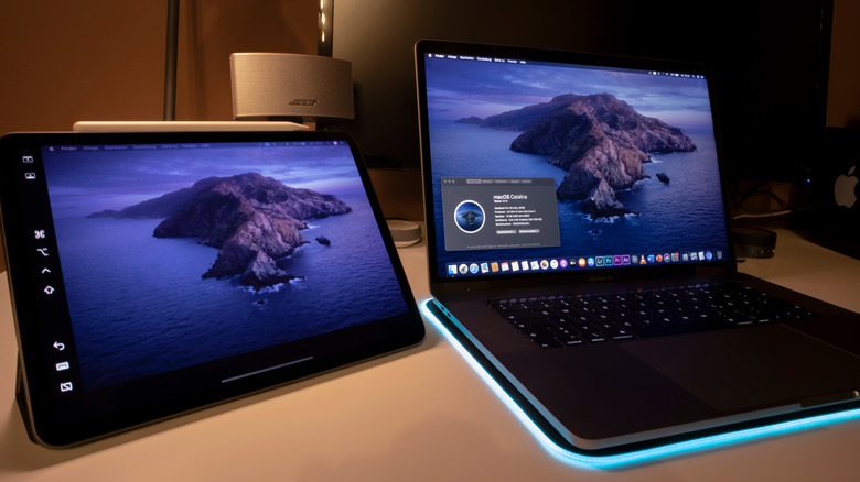 Tablet and laptop side by side on a desk, both displaying the same macOS desktop wallpaper, with a speaker and soft ambient lighting in the background.