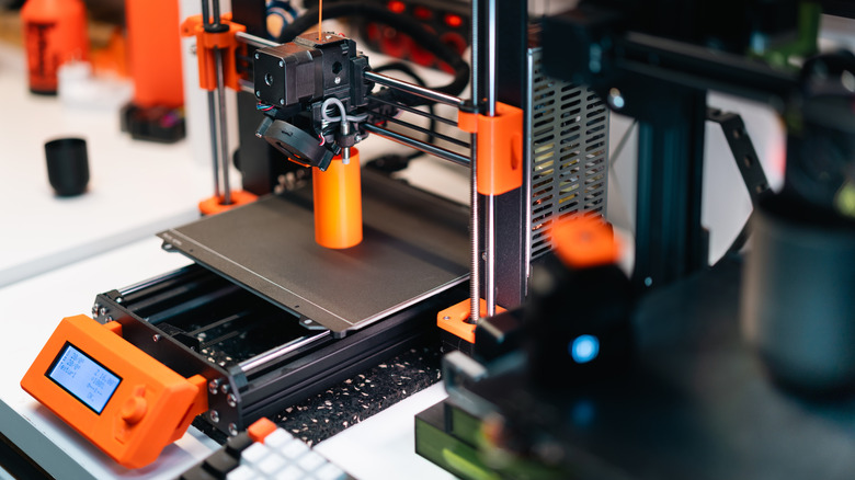 A 3D printer on a white table printing an oranage cylinder