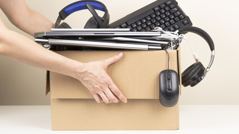 Cardboard box full of and overflowing with electronics like keyboards, mice, headphones, and laptops.