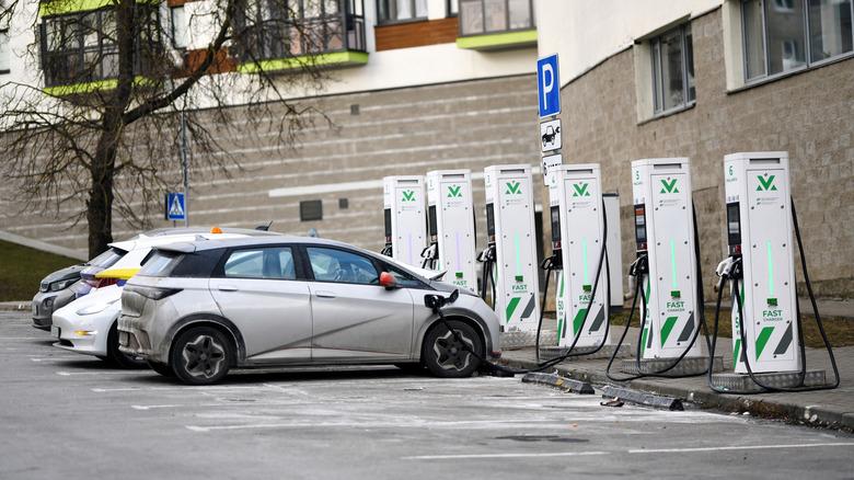 A parking lot containing a row of six EV charging stations, multiple vehicles are taking advantage of the chargers