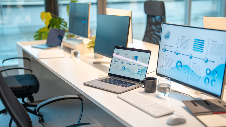 An office space featuring laptops connected to external monitors on a large white desk with cups and peripherals on it.