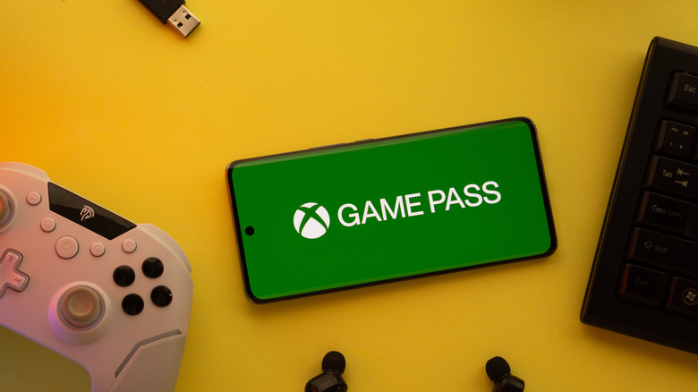 The Xbox Game Pass logo on a smarthpone next to other gadgets on a yellow background