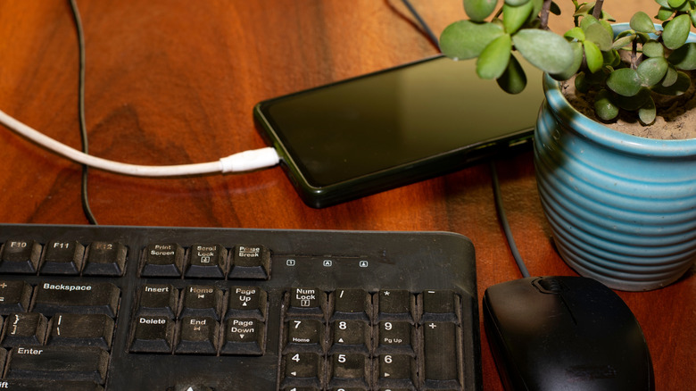 Phone plugged into charging on a work desk alongside a wired keyboard and mouse