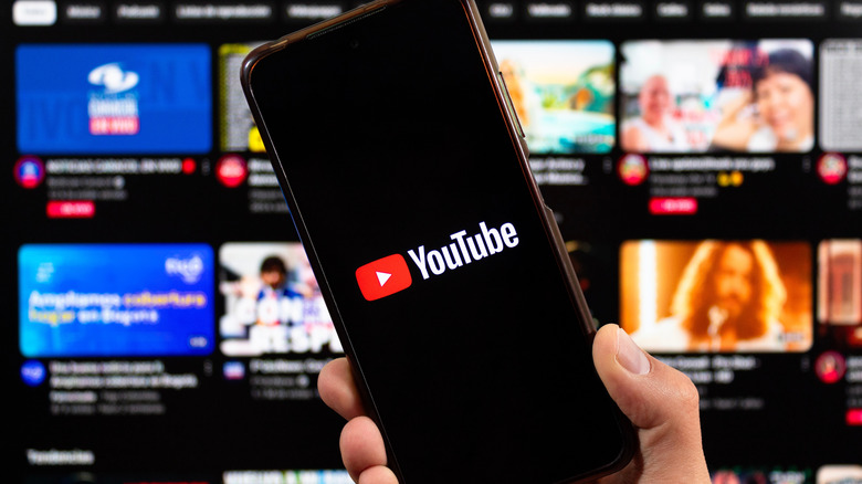 A person holding a phone with the YouTube logo in front of a monitor displaying various YouTube video thumbnails