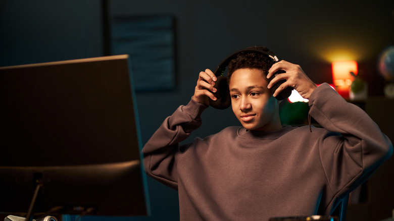A person sitting at a computer monitor wearing a brown sweater placing headphones on their head.