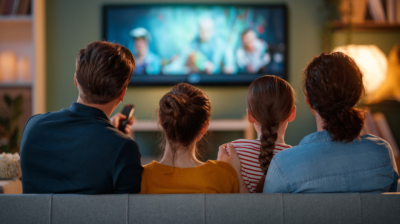 Two children sitting in between two adults, all of them have their backs turned away from the camera as they focus on a television that's hanging from the wall