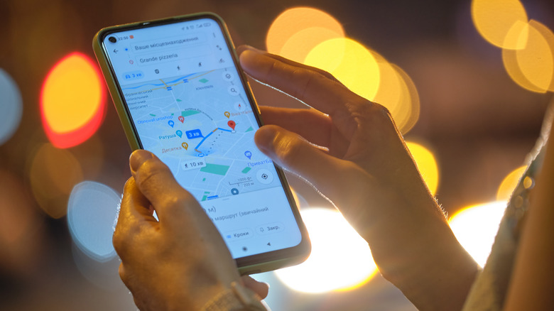 Two hands hold a cellphone in a dark room with lights. The phone shows Google Maps walking directions to a pizza parlor.