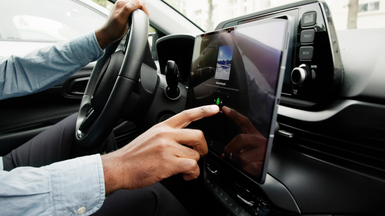 A person driving a car, holding the steering wheel with one hand and tapping the car's touchscreen with the other