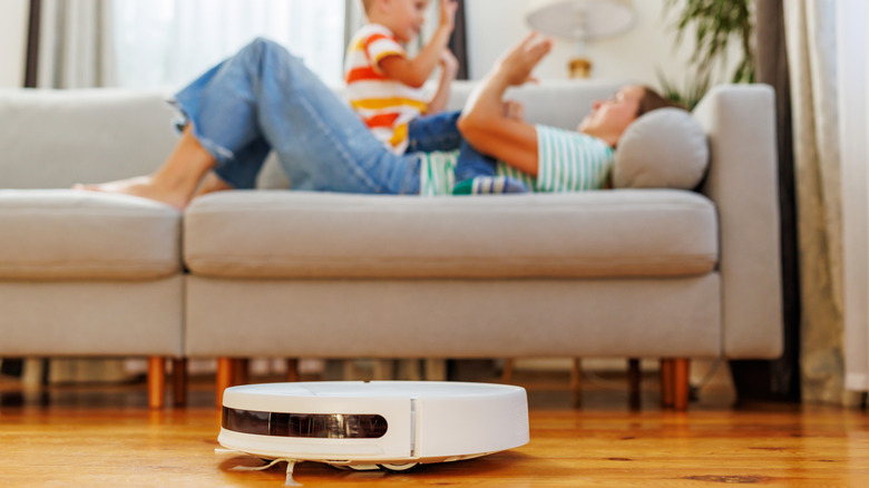 A white robot vacuum cleaner on a hardwood floor with people playing in the background.