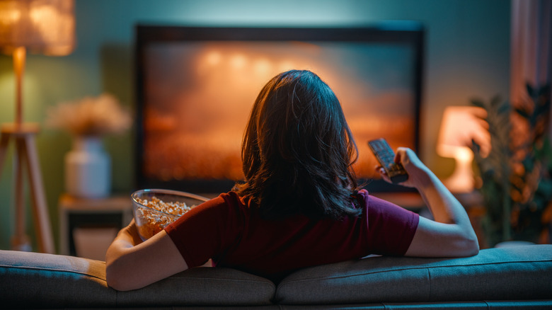 Woman sitting on a couch with popcorn and a remote while watching TV.