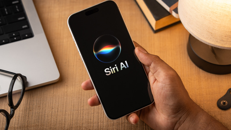 A closeup of a user's right hand holding a smartphone of some kind that is displaying the Siri AI logo, behind the smartphone is a lamp, some books, a pair of glasses, and a silver laptop (likely a MacBook)