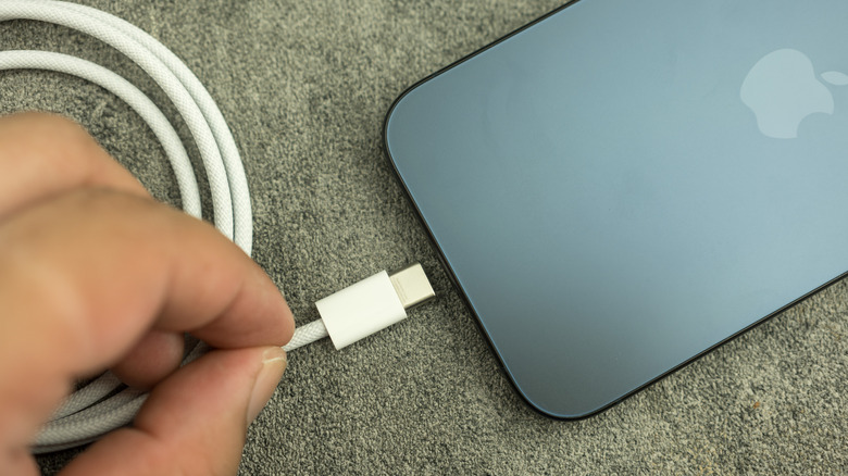 A black iPhone sitting on what appears to be carpet, an individual is plugging a white USB-C charger into the device with their left hand
