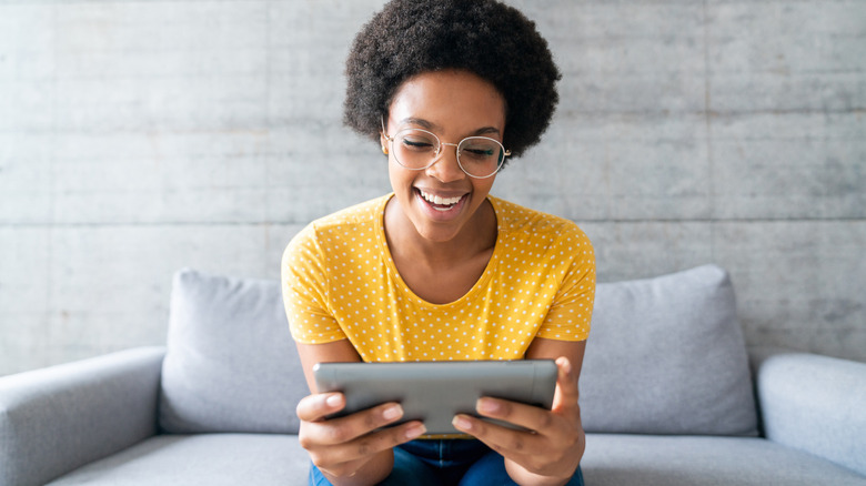 A young woman sitting on her sofa and looking at her tablet with a smile