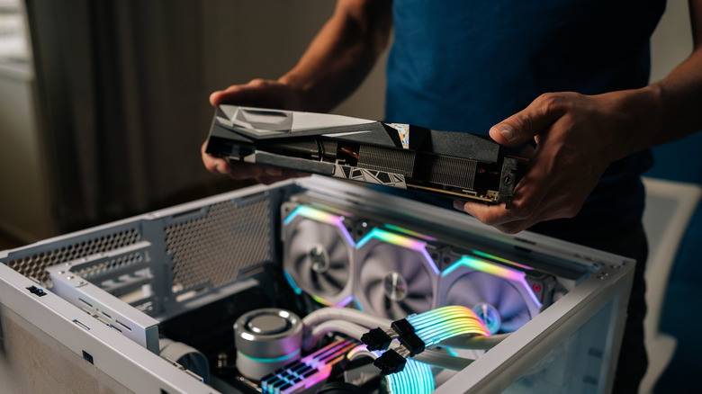 Person holding a graphics card above an RGB-lit desktop PC build.