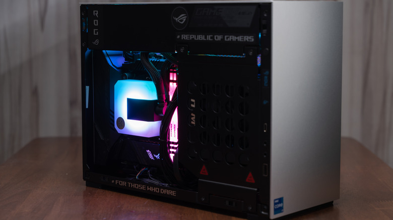 A compact Mini-ITX gaming PC on a wooden table, its glass side showing an Asus ROG graphics card, a liquid CPU cooler lit in cyan, and HyperX RAM glowing pink.
