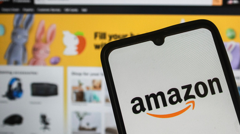 A photograph of the Amazon logo on a smartphone in front of a screen displaying the Amazon homepage.