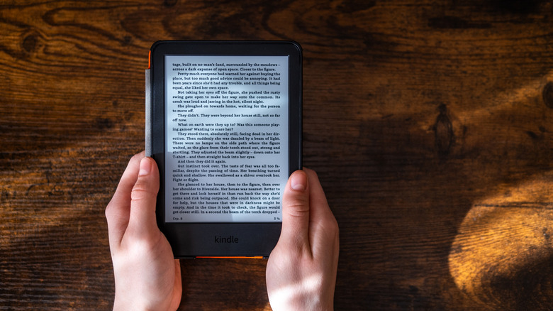 A person holding a Kindle above a wooden table with an e-book opened up.