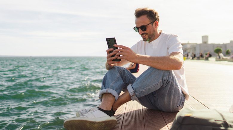 A man sits alongside the ocean and looks at his phone
