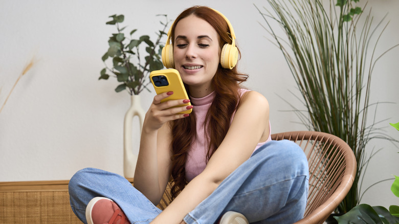 A woman wearing yellow headphones and holding an iPhone with a yellow case while sitting in a wicker chair