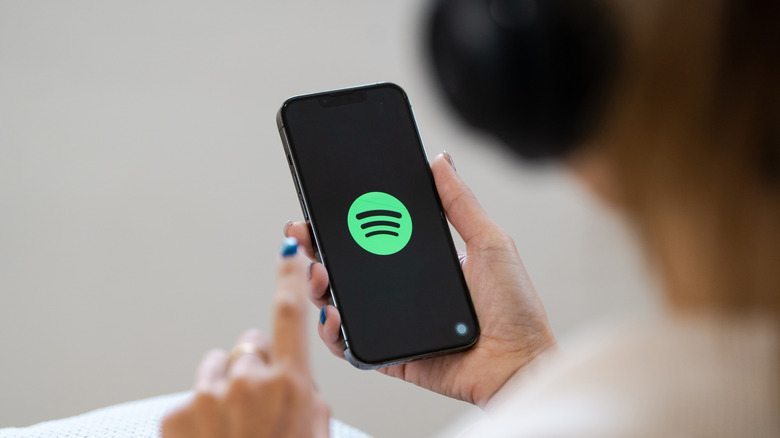 A behind-the-back shot of a woman wearing headphones while hjolding an iPhone displaying the Spotify logo