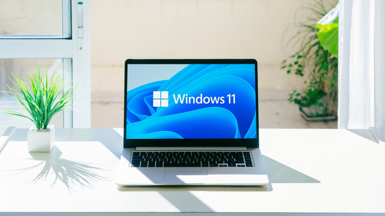 A laptop sitting on a table. On the display of the laptop is th logo for Windows 11.