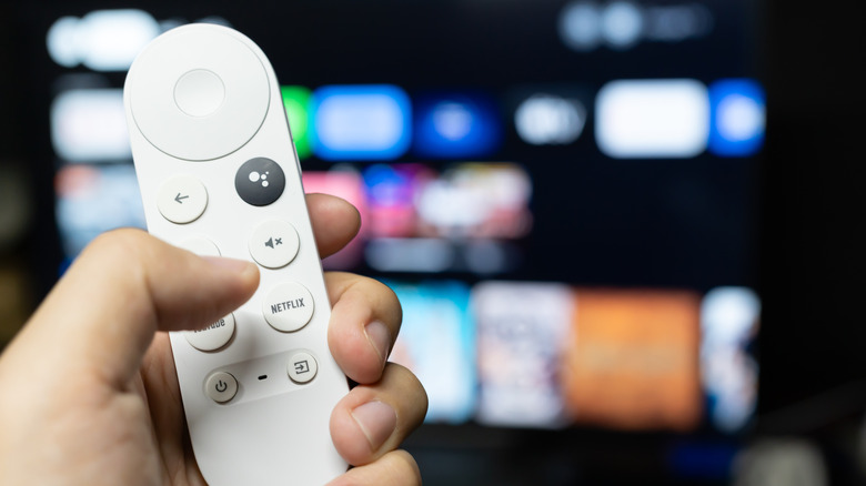 Person holding a google TV remote with a TV screen in the background.