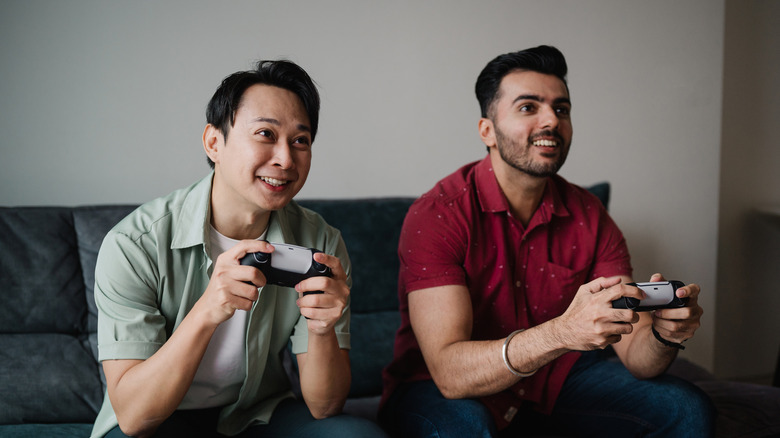 Two men smiling, holding controllers and playing a game on the TV as they sit on a couch