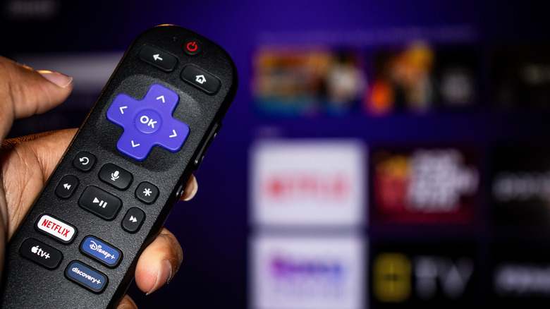 A hand holding a Roku remote in front of a Roku interface
