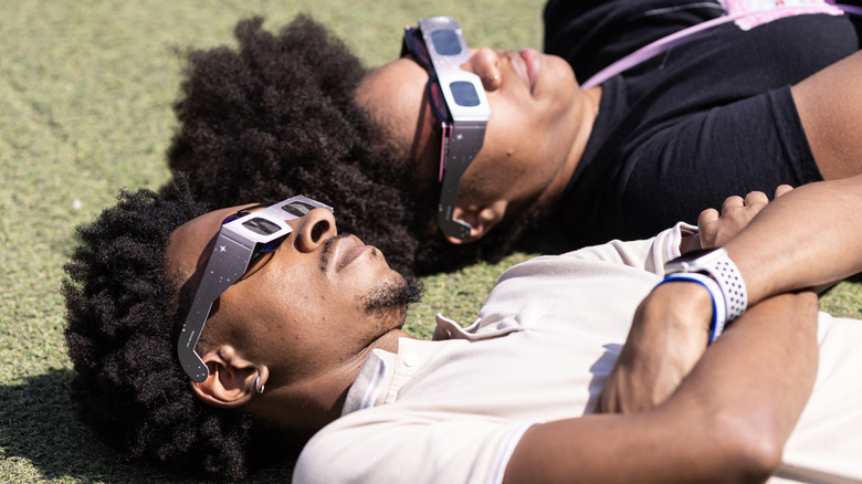 People laying down in the grass looking up at a solar eclipse with eyewear.