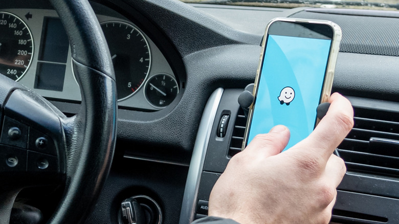 A driver opening the Waze app on a phone mounted on a car dashboard