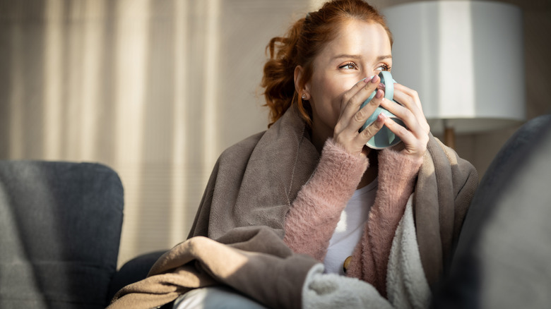 A woman covered in a blanket on a couch drinking coffee.