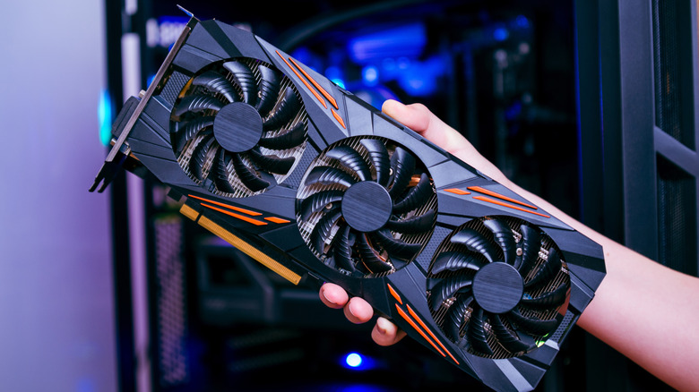A hand holding a black three-fan graphics card with orange accents.