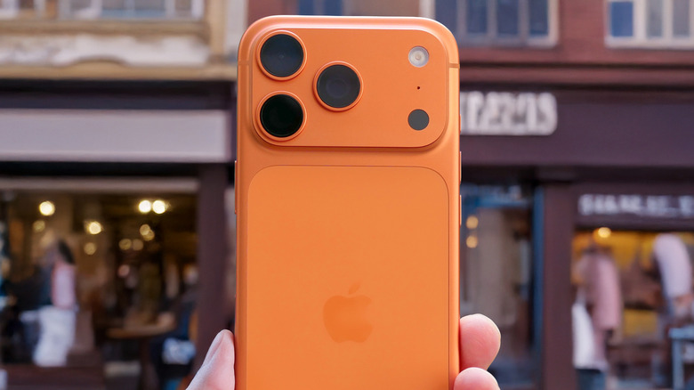 A Cosmic Orange iPhone 17 Pro is held in a user's left hand as they face it toward a line of city buildings