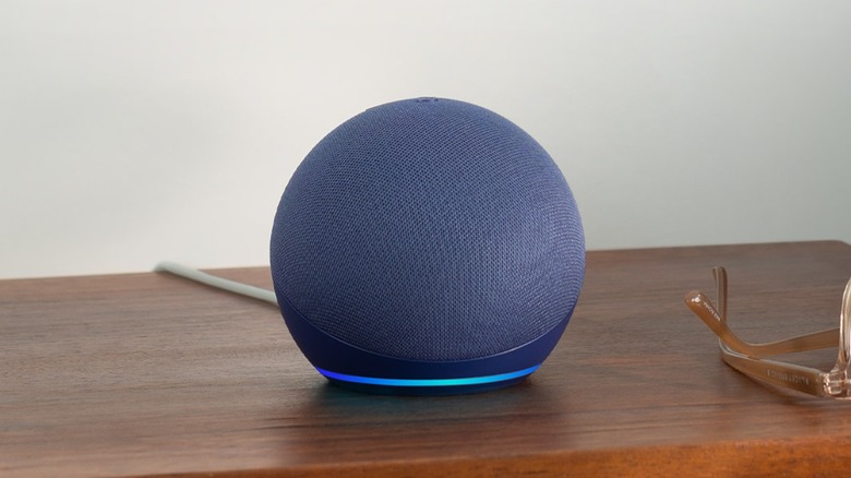 A blue Amazon Echo Dot with its status bar lighting up, the item sits on a desk next to a pair of glasses