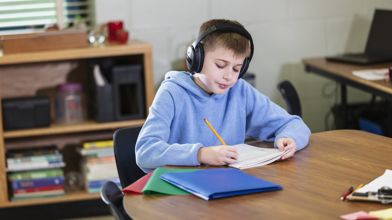 A young boy doing schoolwork at a table while wearing a pair of headphones.