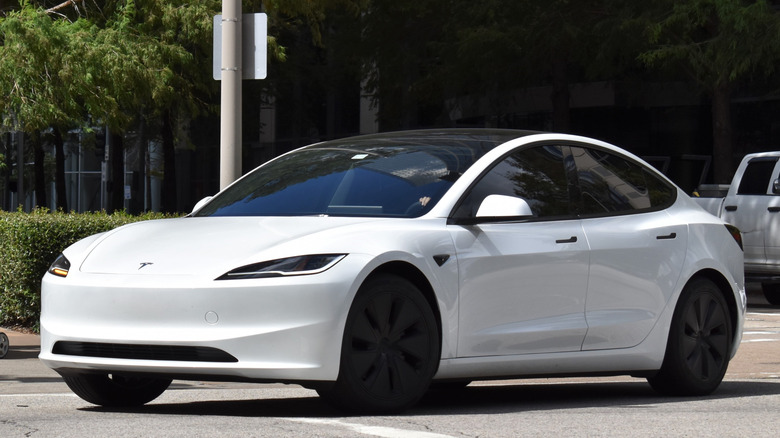 A white Tesla Model 3 as shown from the exterior, the vehicle sits in a parking lot, and some shrubbery can be seen behind it