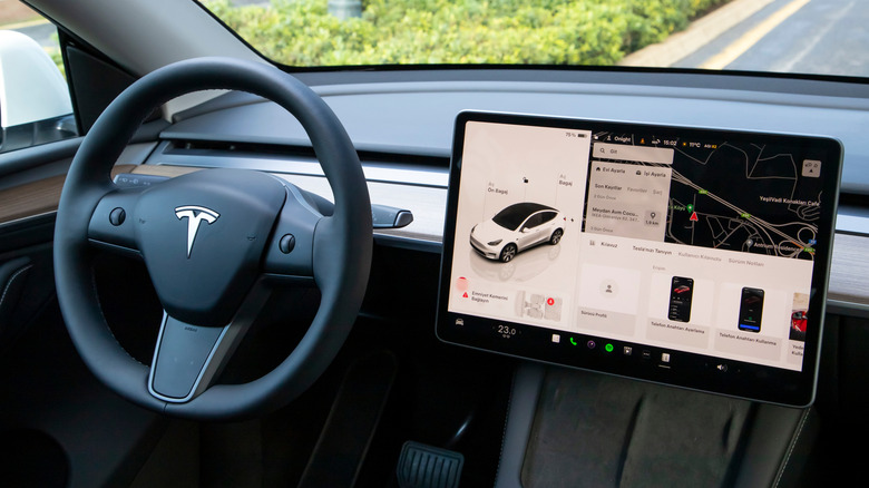 An interior shot of a Tesla vehicle, the image focuses heavily on the steering wheel and the infotainment display, which appears to be showing mapping and smartphone information
