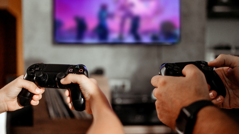 Two people holding PS4 DualShock controllers in front of a blurred, wall-mounted TV in the background.