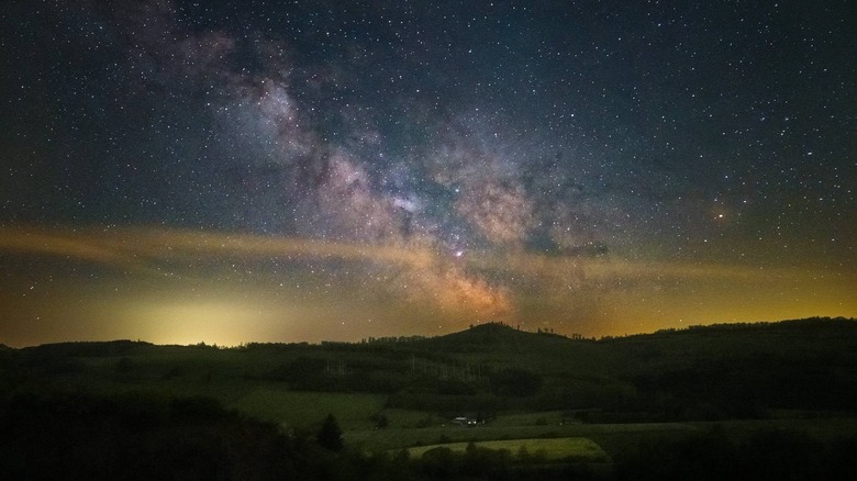 A photo taken by a Google Pixel of a colorful starry night sky over tree-lined rolling hills