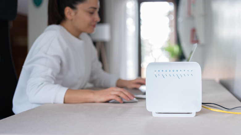 A picture of a router on a table, with a woman in the back using a laptop