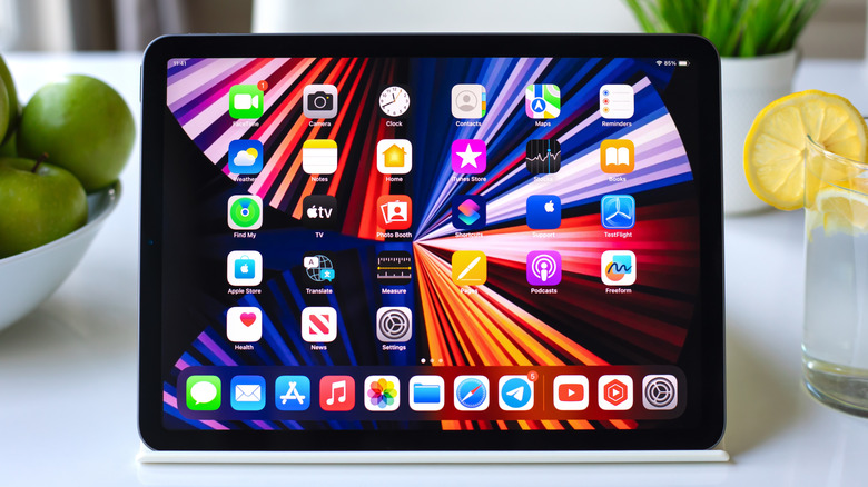 An iPad sitting on a counter inbetween a bowl of green apples and a glass of lemon water, the iPad is displaying the iPadOS 17 Home screen